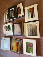 Multiple stacks of photographic prints with matte frames laid out on floor. Images include flowers, trees, sunsets, and landscapes with different colored mattes.