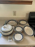 Stoneware dish set arranged on kitchen counter: large serving bowl, covered divided dish, dinner plates, shallow bowls, smaller plates, and saucers all white with blue trim.
