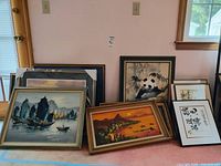 Photo showing multiple framed artworks leaning against a wall including a sailing ships painting, panda artwork, and calligraphy piece.