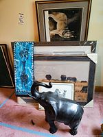 Several framed wildlife prints and photos leaning together including lion, elephants, and zebras, with a blue decorative fabric with animal motifs and a black metal elephant statue in front.