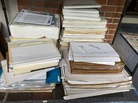 Stacked collection of white and brown storage boxes containing photographic prints, arranged on the ground beside a brick wall.