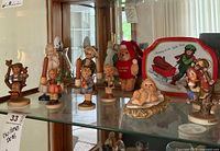 Image showing a collection of Goebel Hummel figurines, a plush bear toy in Christmas attire, and other items inside a glass cabinet.