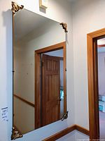 Front and angled views of the rectangular wall mirror mounted on a wall, showing gold ornamental accents on each corner and clear reflective glass surface.