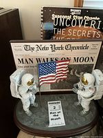 Two astronaut figurines raising US flag on oval base with newspaper front page