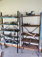 View of two metal shelving units holding multiple model airplanes. The shelving units show rust, wear, and dust. One shelving unit is black metal with metal shelves; the other is dark gray metal with wood or composite shelves.