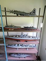 Military-themed model ships and a bomber aircraft displayed on wooden shelves. The aircraft is large with U.S. markings, and the ships vary in size and type, showing detailed military designs. Models show signs of dust and possible minor missing pieces.