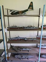 Shelf display showing large military transport plane model on top shelf and multiple gray and red hull military ship models on lower shelves.