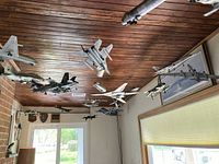 Photo of 18 model airplanes hanging from the ceiling in a room with wooden ceiling, brick wall, and window; airplane models include various military jets and passenger planes in gray, white, and black colors.