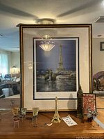 Frontal view of the framed Eiffel Tower print with miniature Eiffel Tower statue, Big Ben statue, metal bicycle sculpture, and shot glasses arranged in front.