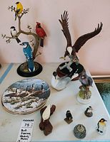 12 porcelain bird figurines and 2 decorative plates shown on a table against a pink wall. Birds include eagle, cardinal, blue jay, small birds, and penguins. Plates feature waterfowl artwork.