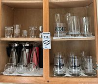 Glassware tumblers and barware stored in wooden cabinet, various sizes and designs visible including Polo and airplane motifs.
