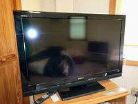 Front angled view of Sony BRAVIA 42-inch LCD TV. Remote control placed on wooden stand beneath TV. Minor dust visible on TV surface.