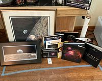 Collection of framed space-themed prints and multiple space-related books arranged together on a wooden floor with bookshelf background