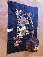 Photo showing the textile fabric laid flat with embroidered elephants and figures in traditional attire along with the circular decorative bamboo hat placed on the corner.