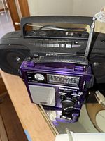Close-up image of a vintage purple Grundig receiver with tuning dials, large speaker grille, and antenna, in front of a black Sony boombox.