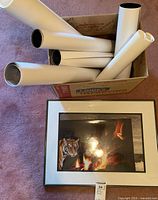 Photo showing multiple rolled artistic prints inside a box and one visible framed print laid flat, depicting a tiger near fire.