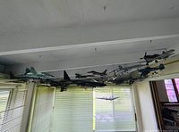 View of multiple vintage military model airplanes suspended from ceiling in a room with window blinds.