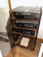 Full view of wooden stereo cabinet with glass door open showing three stereo components inside and a manual on bottom shelf.