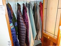 Side view of seven medium jackets showing various colors including blue, green and purple, hung on gold wire hangers, showing Antarctica patches