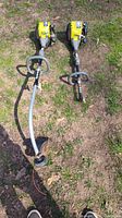 Two yellow Ryobi gas weed wackers placed on grass showing full length and handle design. One weed wacker is missing part of the trimmer end.