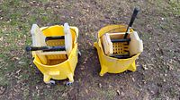 Two yellow wet floor mop buckets with wringers shown outdoors on grass with dirt and leaves. Both have wheels and caution signs visible.
