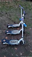 Three Razor Power Core E100 scooters lying on grass outdoors with visible wear and dirt. Purple and blue handle grips, black foot decks with Razor logos.