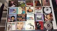 40 assorted music CDs arranged in rows on a table, showing a variety of cover art with portraits, text, and graphics including artists like Billy Joel and an album titled 'Through the eyes of Julie'.
