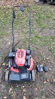 Full view of red Craftsman gas lawn mower on grass with handle upright, showing included battery and charger on ground nearby.