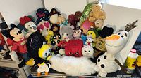 Wide view of a group of 21 assorted plush toys including Disney characters (Mickey, Minnie, Olaf), novelty plush (Hershey's chocolate bar), multiple animals and other soft toys in various sizes and colors arranged on a surface.