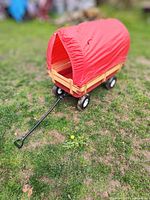 Side angle photo showing overall wagon with red fabric canopy cover and pull handle, sitting on grass.