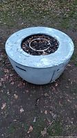 Large round concrete gas fire pit showing top and side views with weathered surface and metal gas ring burner filled with rocks.