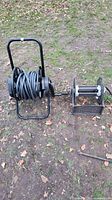 Two hose reels side by side outdoors on grass; left is tabletop style reel, right is mobile style reel with wheels and handle, both have gray hoses wound.