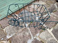 Green metal wheelbarrow-shaped plant holder lying on stone patio, side view showing decorative curls on sides and open grid base.