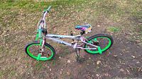 Side angle view of the BMX bike showing the pinkish handlebars and green wheels, green pedal pegs, and colorful seat placed on grass with dry leaves.