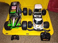 Two RC cars with remote controllers placed on yellow tray on carpet floor, displaying their top views and controllers in front.