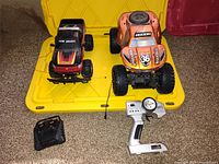 Two remote control cars on yellow plastic container with their remote controls. One car is red and black with New Bright branding, the other is orange with Rockzilla MaxTruck branding.