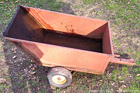 Angle view of small red metal dump trailer with two tires on grass, showing interior and hitch attachment.