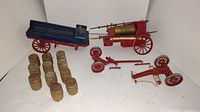 Photo showing disassembled vintage toy fire engine parts including the hose reel with brass drum, two wheel axles, a blue wagon, and 12 small wooden barrels.