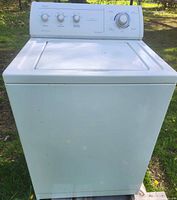 White Whirlpool top load washing machine outside on grass with control knobs and top lid closed, showing the full front and top view.