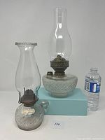 Two antique oil lamps, one with patterned sunburst glass and ruffled chimney, the other with ornate geometric patterned glass and clear chimney, shown with a water bottle for scale.