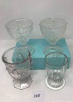 Four antique pressed glass drinking glasses displayed on light blue box against white background, showing two similar stemmed glasses with floral patterns, one hexagonal base glass with bubbles, and one smaller ribbed glass.