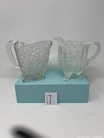 Two clear glass pitchers displayed side by side. Left pitcher has Daisy and Button pattern with a triangular shape and clear handle; right pitcher is opalescent with floral ribbed pattern and three feet.
