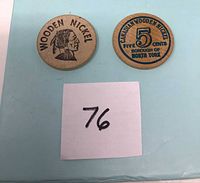 Two wooden nickels displayed on a light blue surface with a numbered sticker '76'. One shows a profile of a native figure with 'WOODEN NICKEL' text; the other shows denomination and North York references.