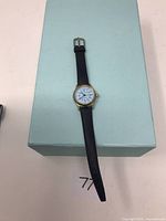 Full view of the gold-tone analog watch with black leather band laid on a light blue box.