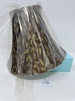 Side angle view of the beige and brown fabric lampshade wrapped in clear plastic.