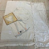 Photo showing multiple white linens including a folded embroidered cloth, a lace-bordered runner, a scalloped-edge doily, and packaged handkerchief.