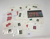 Wide view of multiple German WWII era stamps mounted on paper and some loose, showing various red, green and dark colored stamps.