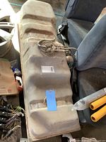 Older GM truck fuel tank placed on the floor beside vehicle seats, showing the fuel pump assembly and attached hoses from the top.