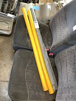 Three yellow tubular handrail covers placed on a car seat for scale and visibility, two unwrapped and one wrapped in plastic.
