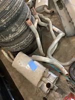 Photo showing two large mufflers, several bent exhaust pipes including the H-pipe, resting on the floor near stacked tires.